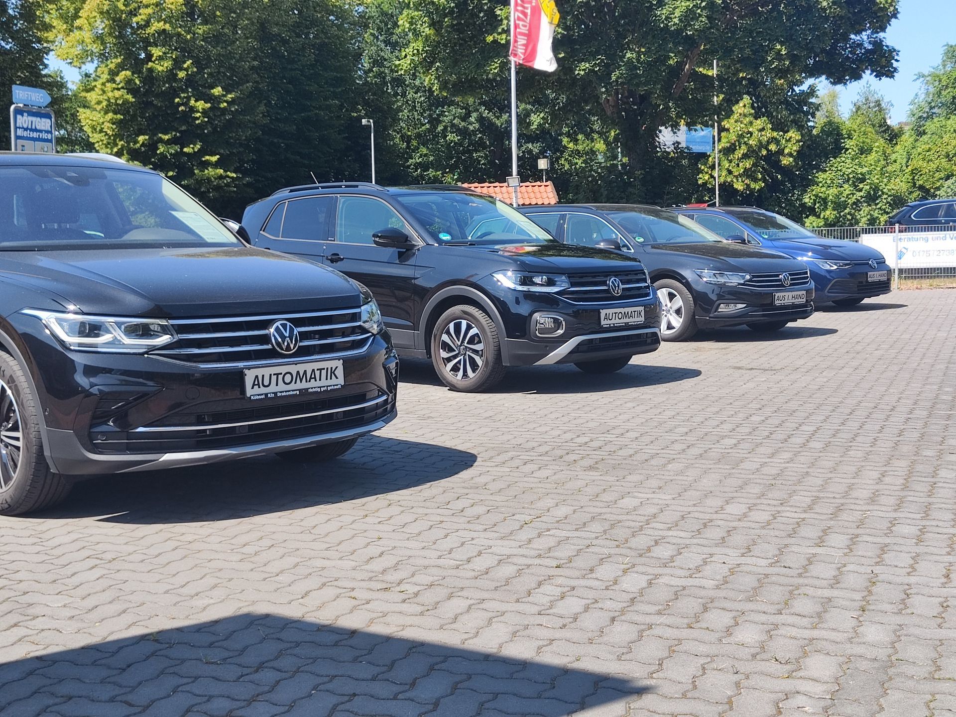 Eine Reihe dunkler Geländewagen steht auf einem sonnigen, gepflasterten Parkplatz neben Bäumen und einem Fahnenmast.