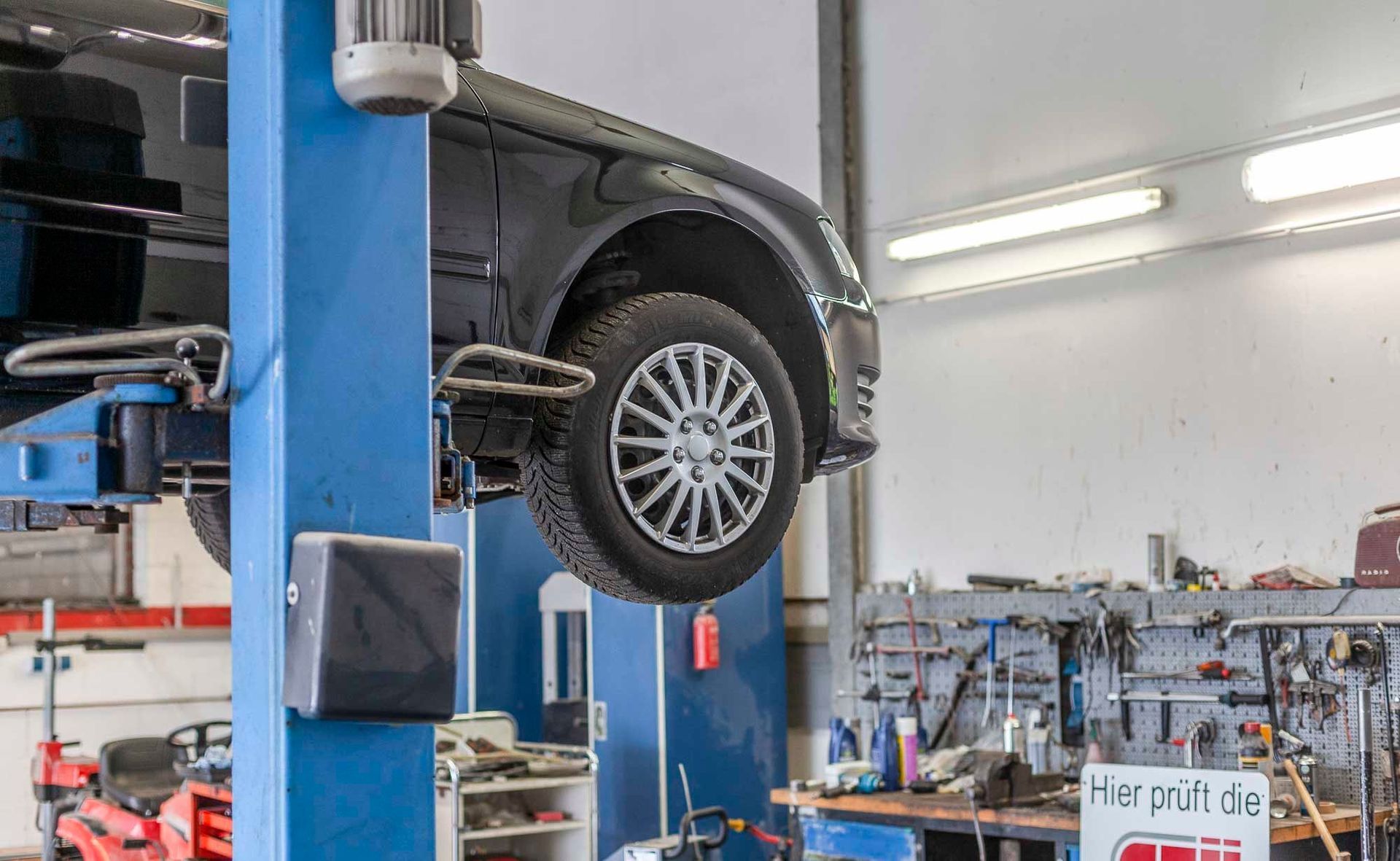 Ein schwarzes Auto wird in einer Autowerkstatt auf einer blauen Hebebühne angehoben; im Hintergrund sind Werkstattwerkzeuge zu sehen.