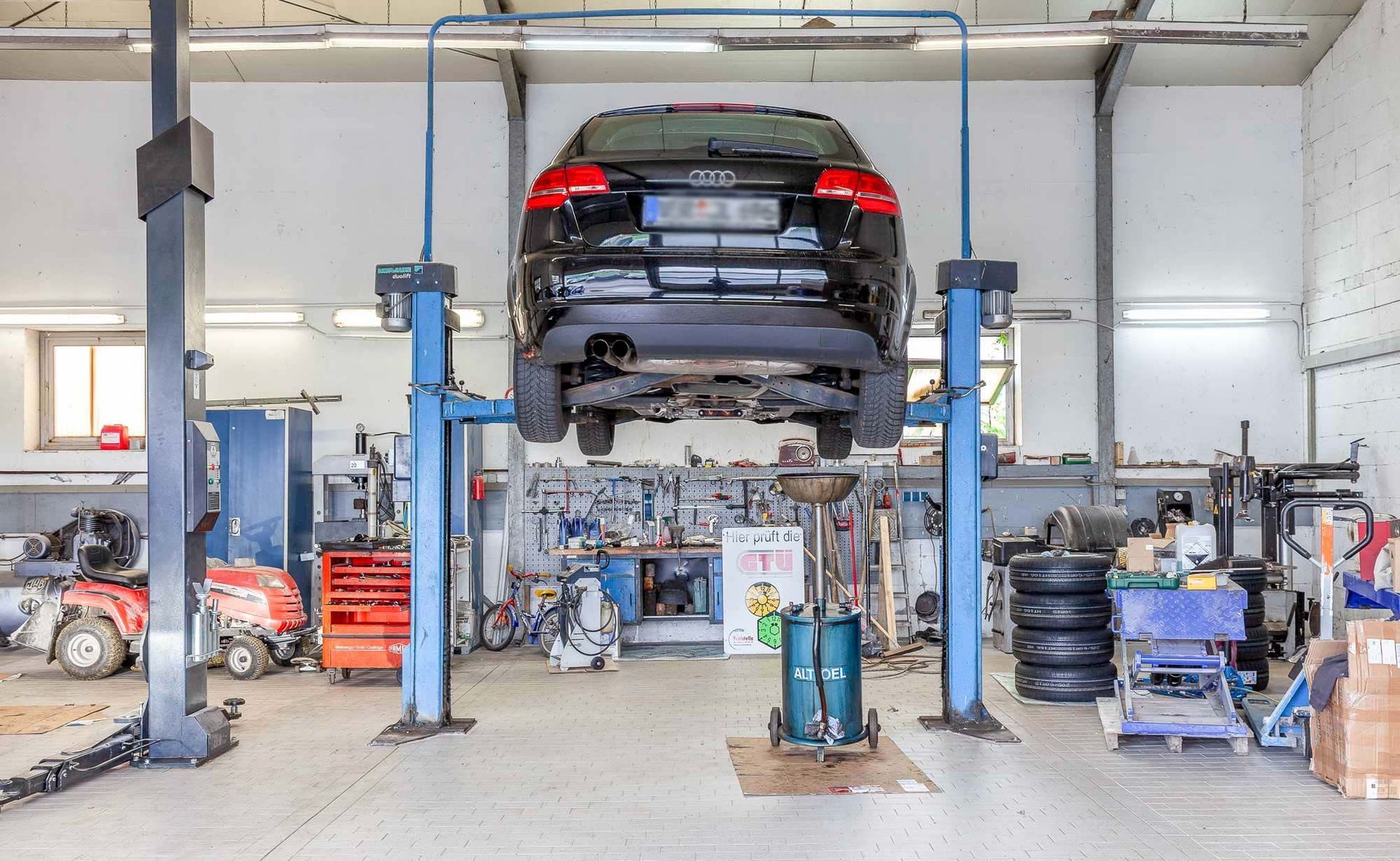 Ein Auto wird in einer Autowerkstatt auf einer Hebebühne angehoben; Werkzeuge und Reifen liegen drumherum.