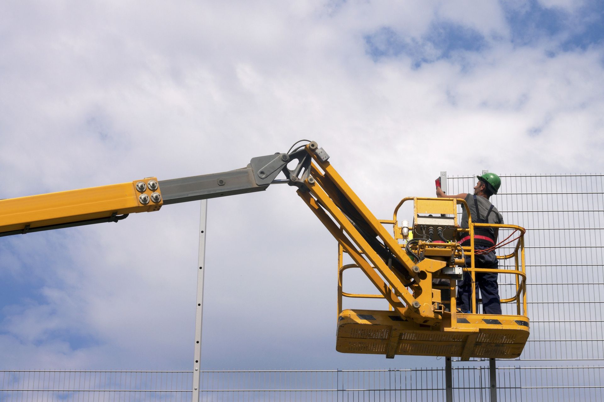Obreros de la construcción usan una grúa para levantar una viga de hormigón. Un trabajador está de pie en una plataforma elevadora amarilla, y el cielo es de un azul brillante.