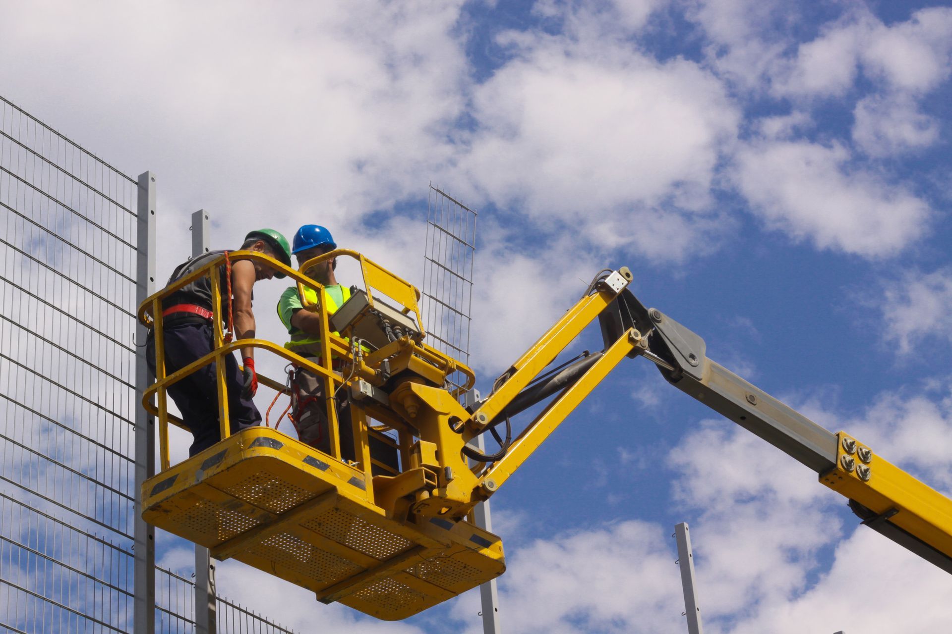 Dos trabajadores de la construcción en un ascensor amarillo trabajando en una cerca de alambre contra un cielo azul con nubes.