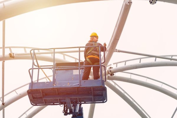 Un trabajador de la construcción con casco amarillo y arnés en un elevador, inspeccionando una estructura metálica grande y curva.