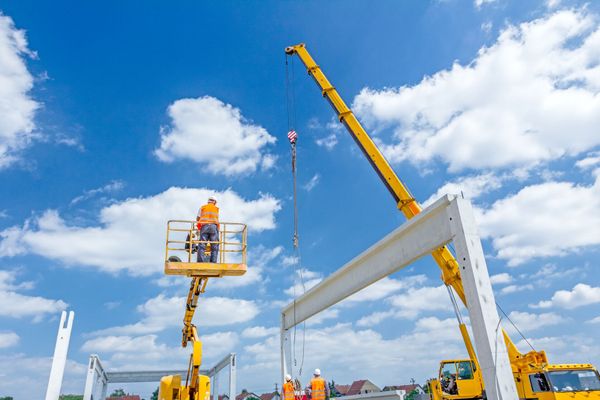Obreros de la construcción usan una grúa para levantar una viga de hormigón. Un trabajador está de pie en una plataforma elevadora amarilla, y el cielo es de un azul brillante.