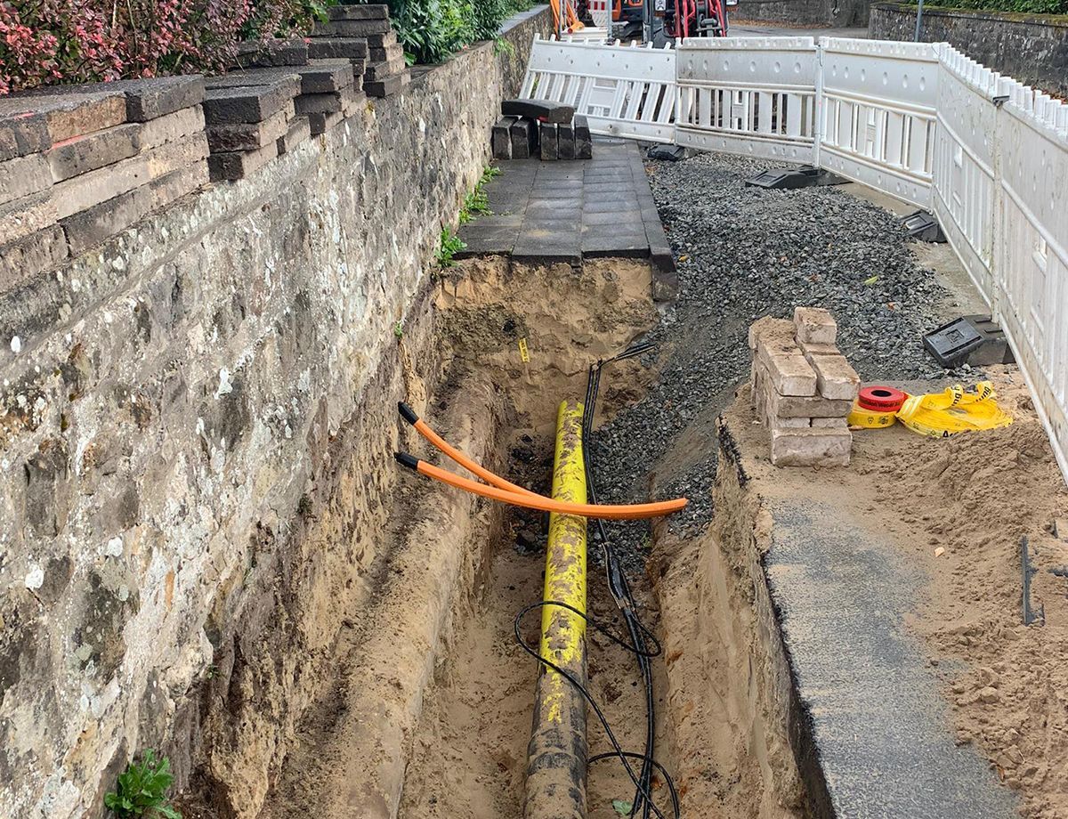 Neben einer Steinmauer wird ein gelbes Rohr im Boden verlegt.
