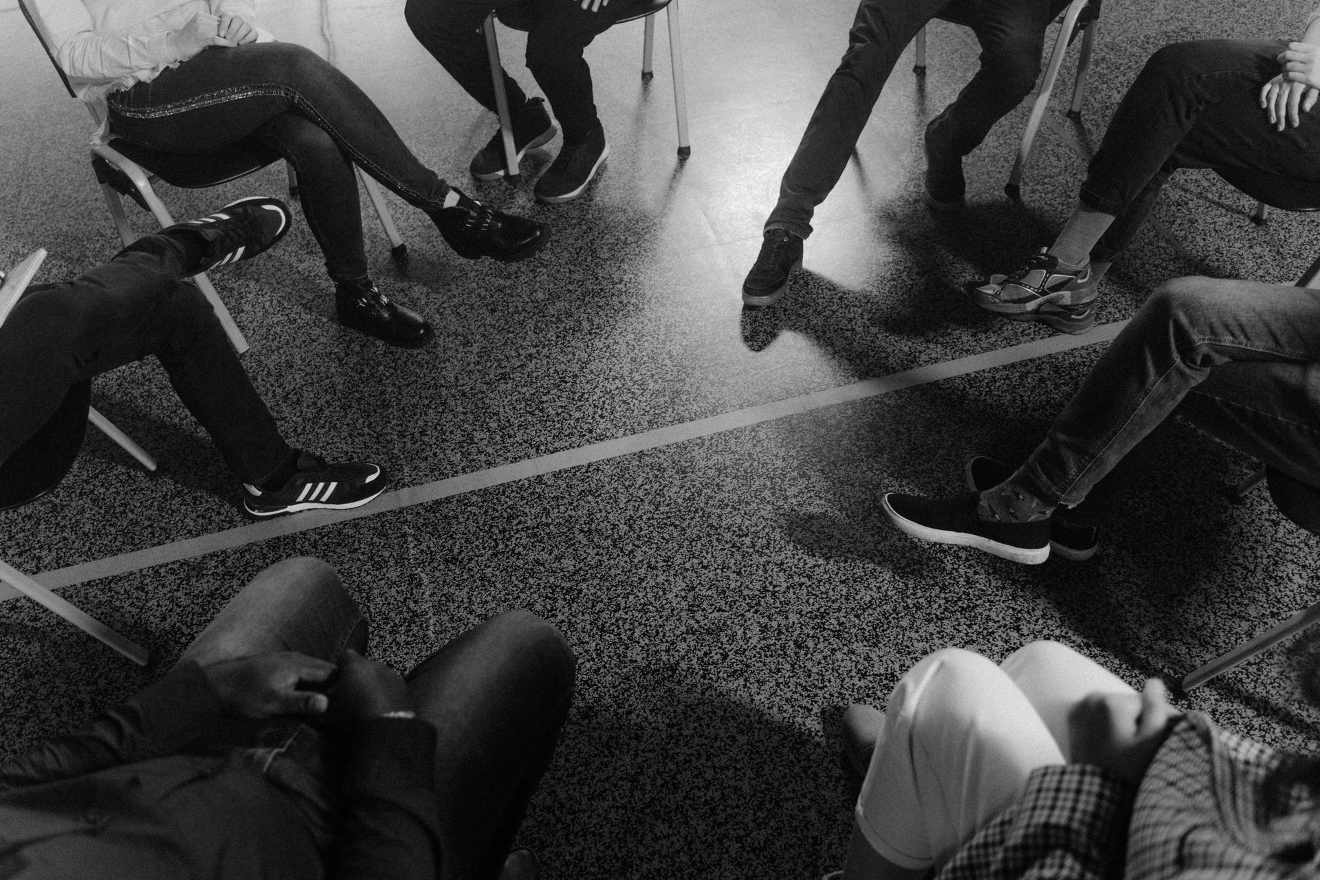 Personas en círculo, sentadas en sillas. Piernas cruzadas, zapatillas blancas y negras visibles. El suelo es gris.