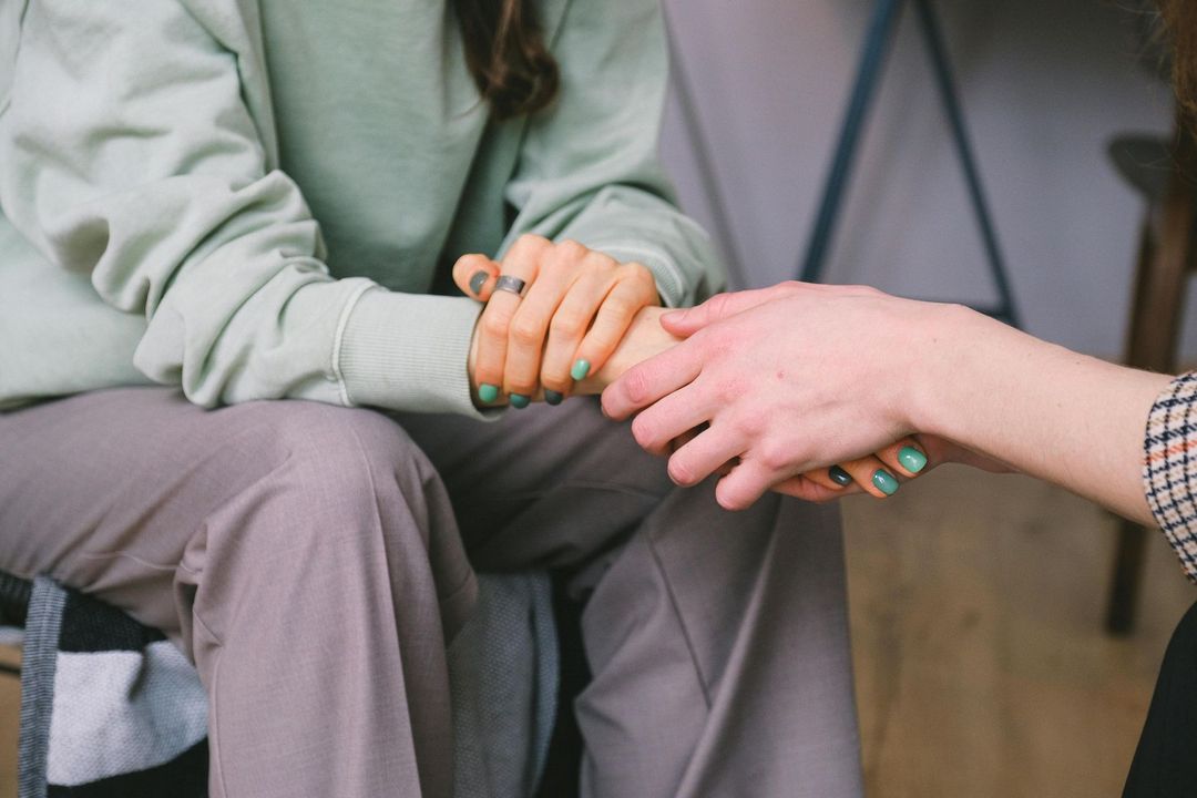 Dos personas tomadas de la mano: consuelo y apoyo. Una con sudadera gris, la otra con camisa a cuadros, en un ambiente interior.