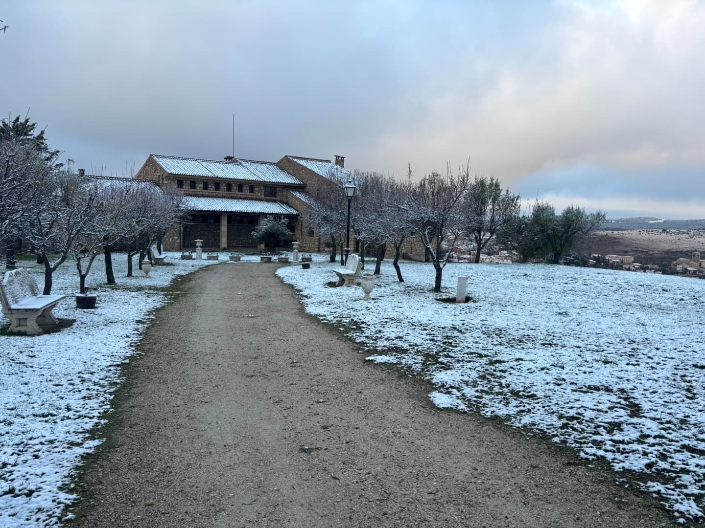 Un sendero nevado conduce a un edificio con el cielo nublado. Árboles y bancos bordean el camino.