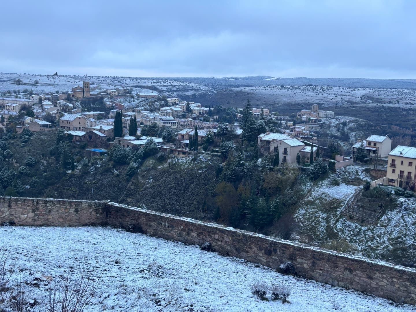 Paisaje de pueblo nevado con muro de piedra en primer plano. Casas y árboles cubiertos de nieve bajo un cielo nublado.