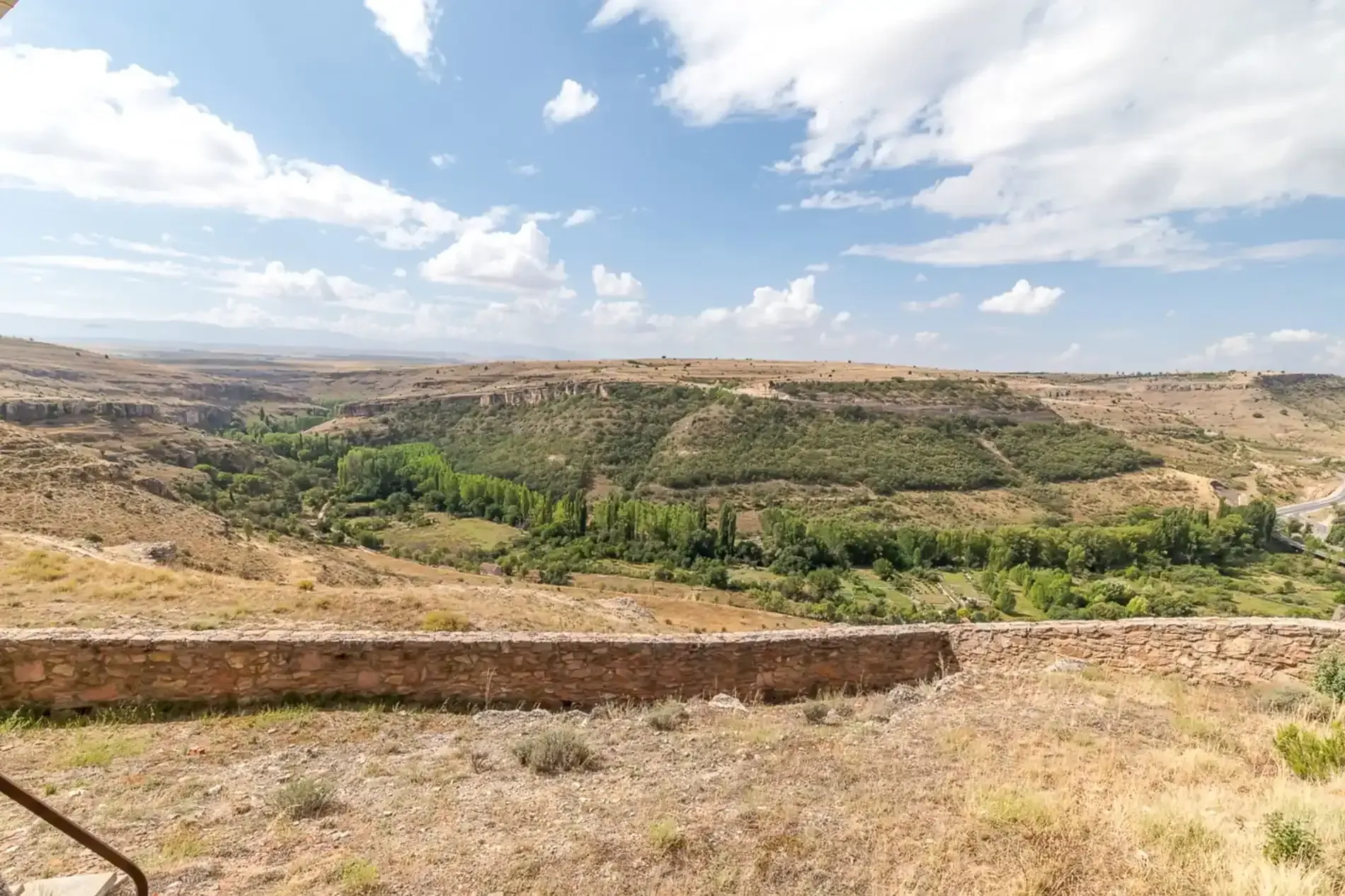 Vista de un cañón con un primer plano seco y herboso y árboles verdes en el valle de abajo; cielo azul con nubes.