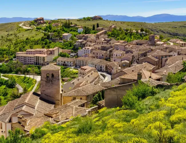 Vista de una ciudad europea histórica con techos de tejas, una torre de iglesia de piedra y colinas verdes al fondo.