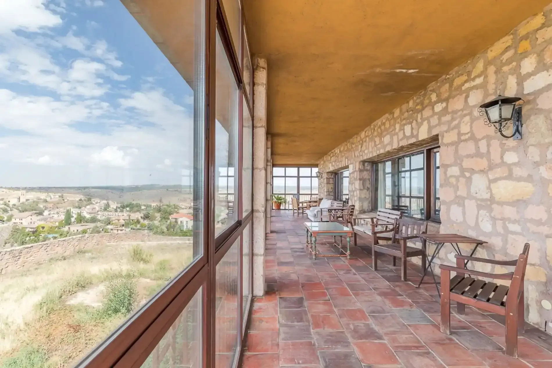 Patio al aire libre con paredes de piedra, piso de baldosas rojas y grandes ventanales con vista a un valle pintoresco.