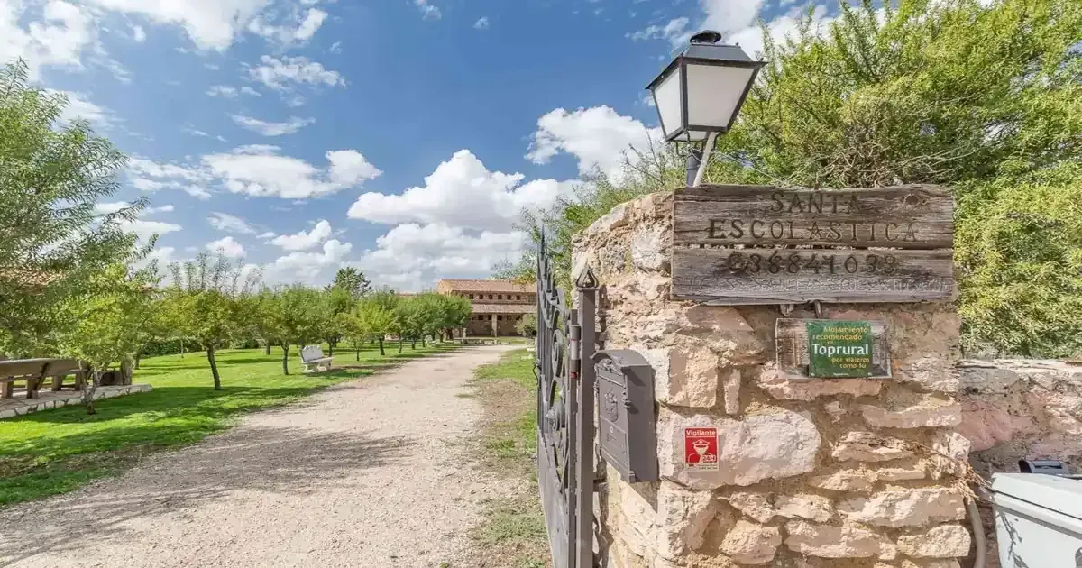 Puerta de piedra con un cartel; un largo camino conduce a un edificio de ladrillo bajo un cielo soleado.