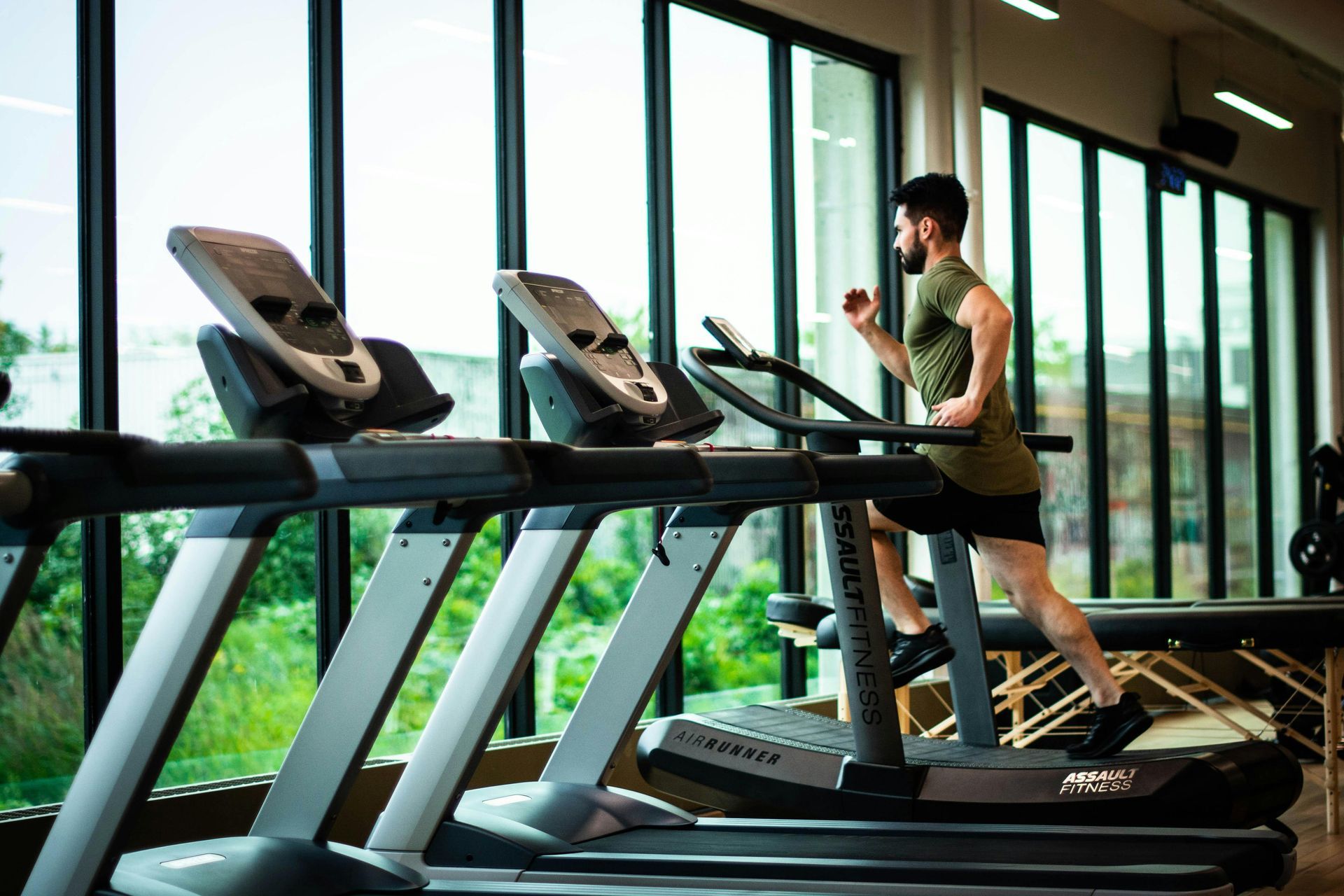 Ein Mann läuft auf einem Laufband in einem Fitnessstudio; im Hintergrund sind große Fenster zu sehen.