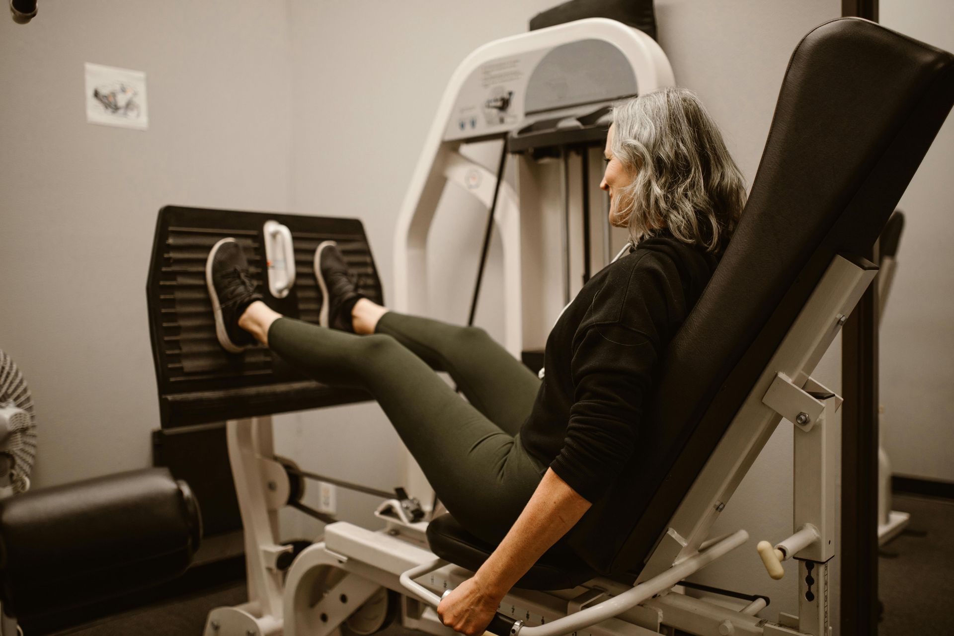 Frau an einer Beinpresse im Fitnessstudio, Beine ausgestreckt, graues Haar.