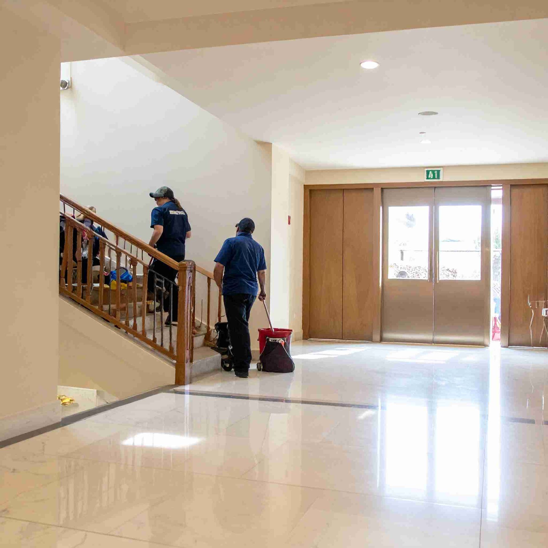 Dos conserjes limpiando un pasillo bien iluminado con escaleras y puertas de salida.