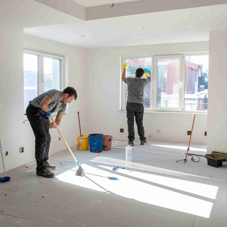 Dos trabajadores limpiando una habitación: uno fregando, el otro limpiando ventanas. Habitación luminosa, suelo de hormigón.