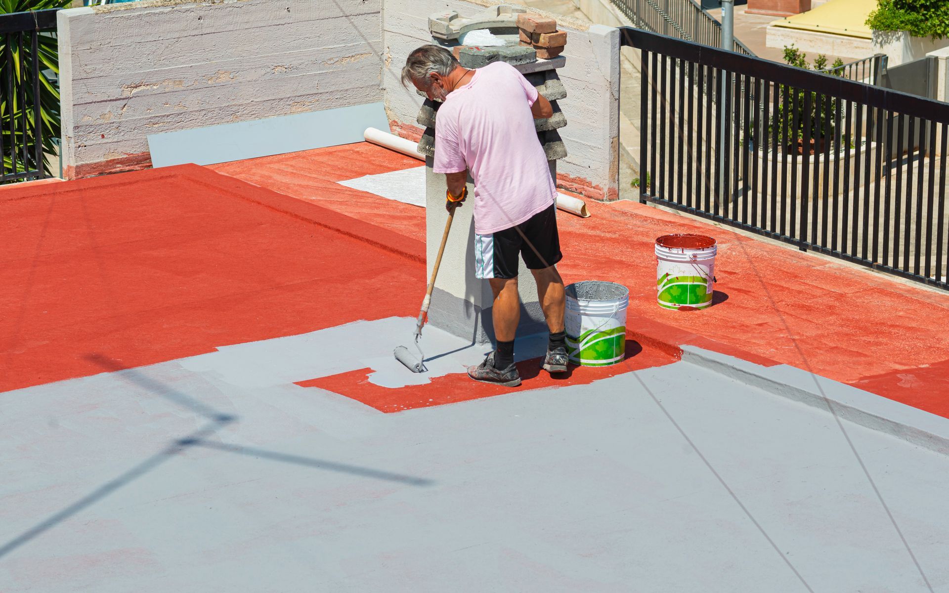 Des ouvriers peignent un toit-terrasse ; l’un étale un revêtement au rouleau, près de seaux et d’outils.
