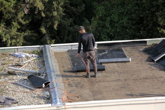 Ouvrier posant une membrane d’étanchéité sur un toit plat, avec du gravier et des arbres en arrière-plan.