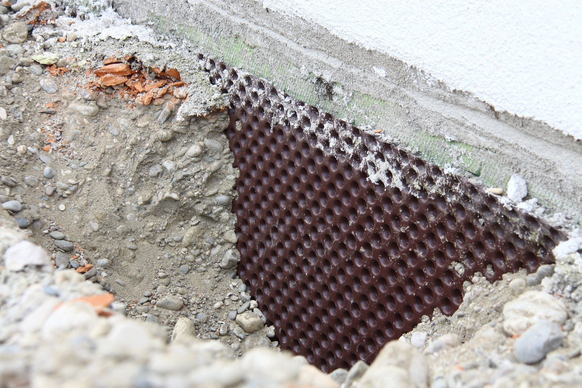 Gros plan sur l’angle d’une fondation en béton, avec une membrane d’étanchéité en maille apparente et des bords irréguliers de gravats.