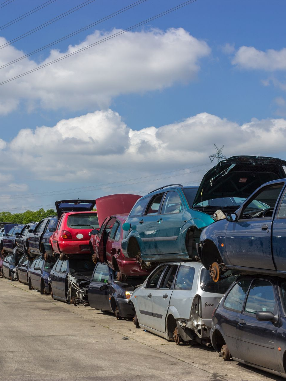 Des épaves de voitures empilées dans une casse, sous un ciel bleu parsemé de nuages.