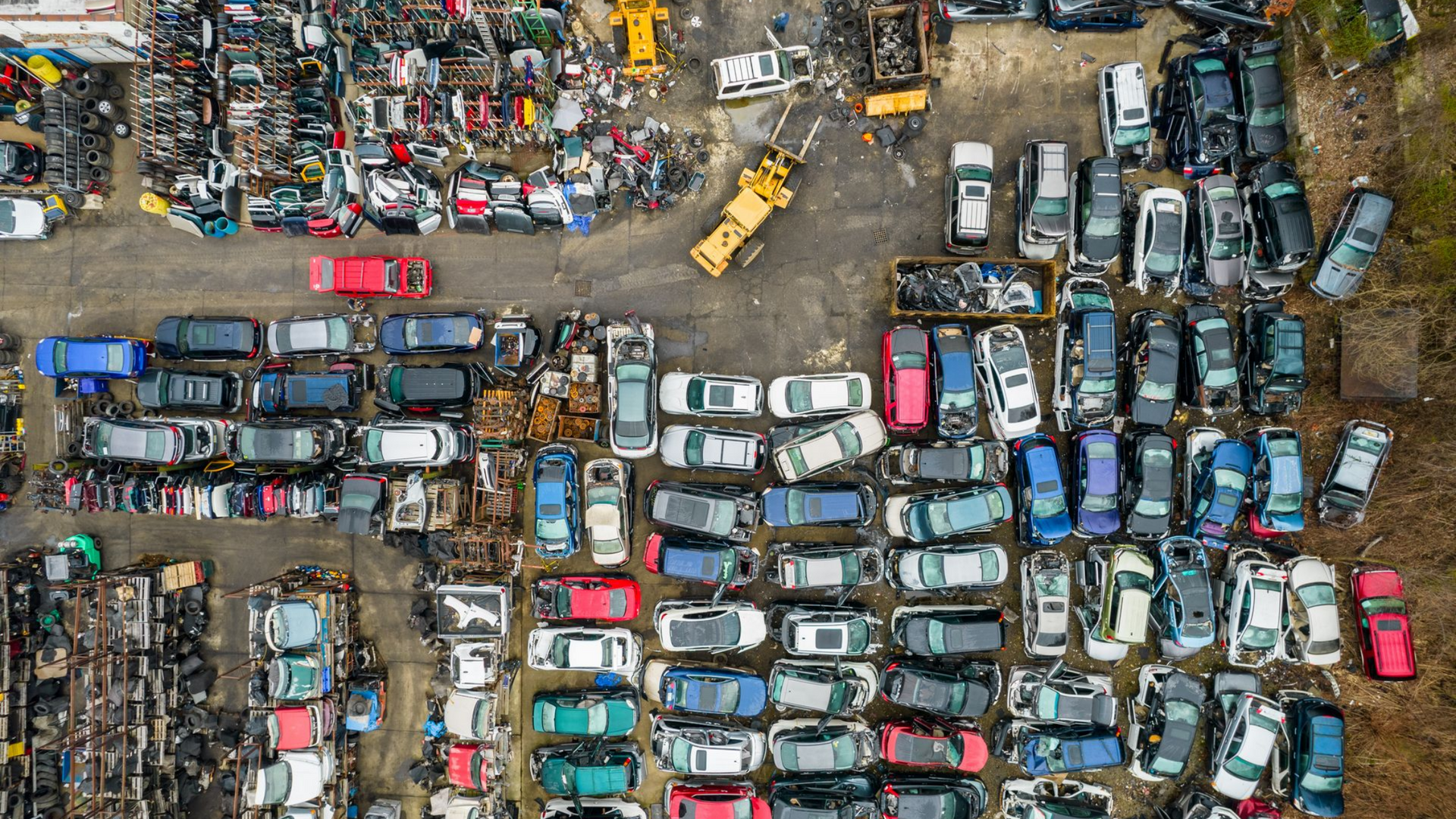 Vue aérienne d'une casse remplie de rangées de voitures accidentées ; un engin de chantier jaune se déplace parmi elles.