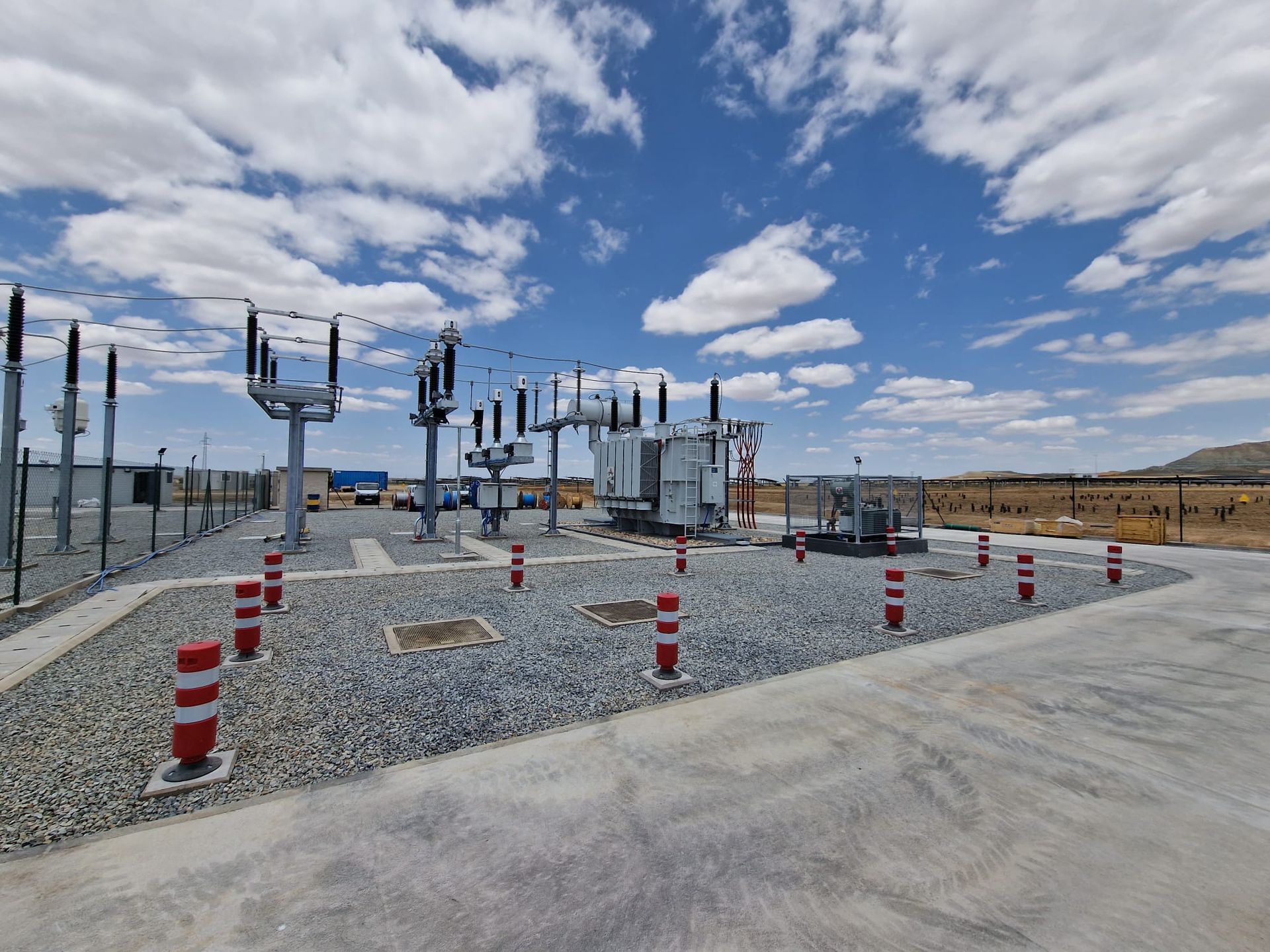 Una subestación eléctrica en un patio de grava bajo un cielo parcialmente nublado. Bolardos rojos y blancos.