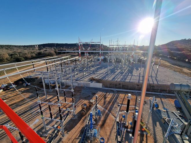 Vista aérea de una subestación eléctrica con equipos plateados y azules bajo un sol brillante.