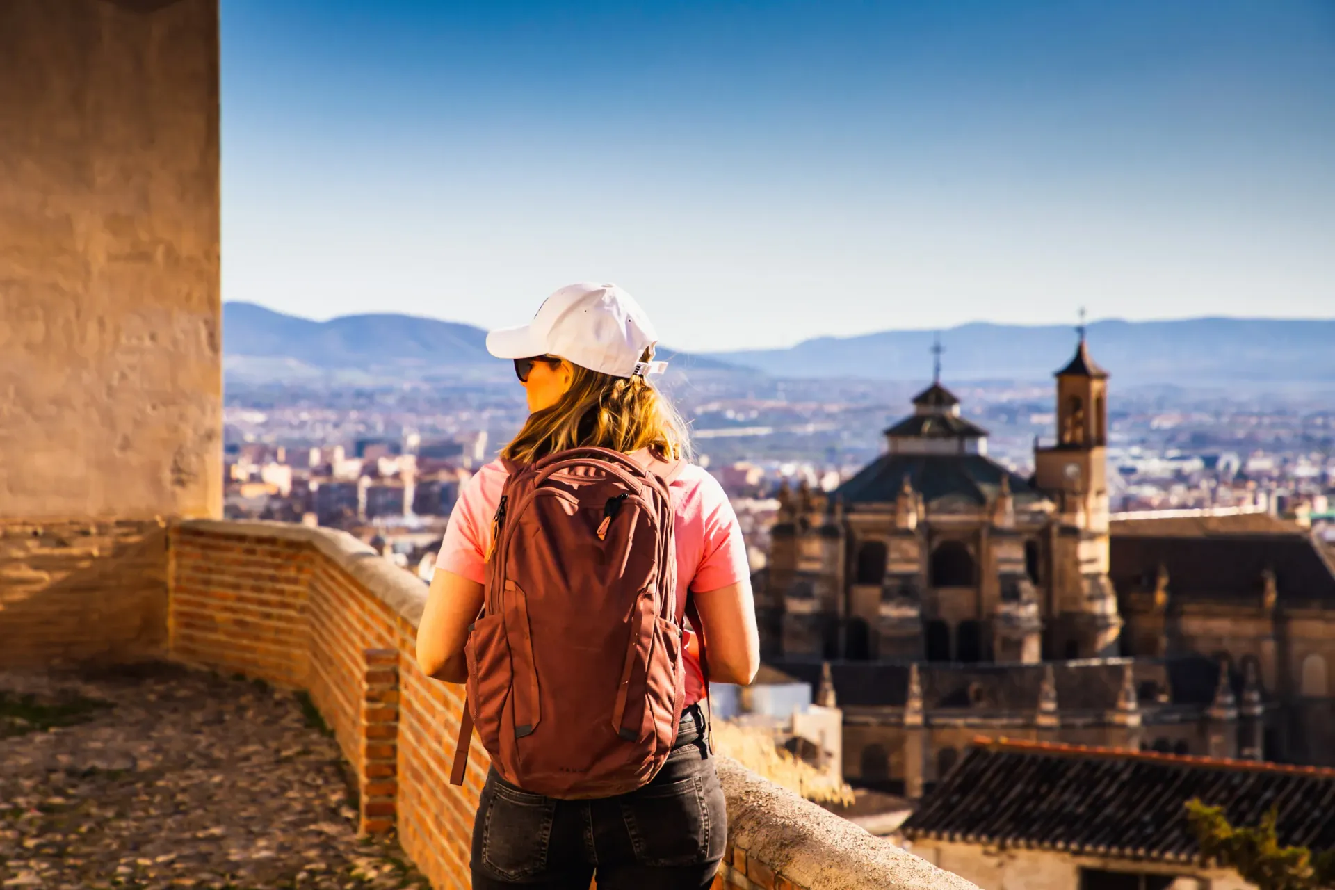 Una mujer con mochila contempla la ciudad y la iglesia desde un muro de piedra.