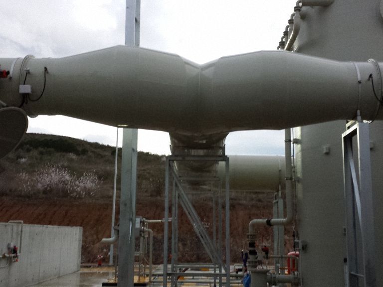Un gran conducto eléctrico gris en un entorno exterior, conectado a un transformador. Al fondo se ven estructuras.