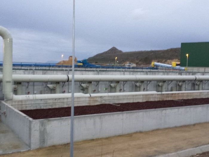 Una planta de tratamiento de agua con depósitos y tuberías de hormigón con una ladera de fondo bajo un cielo nublado.