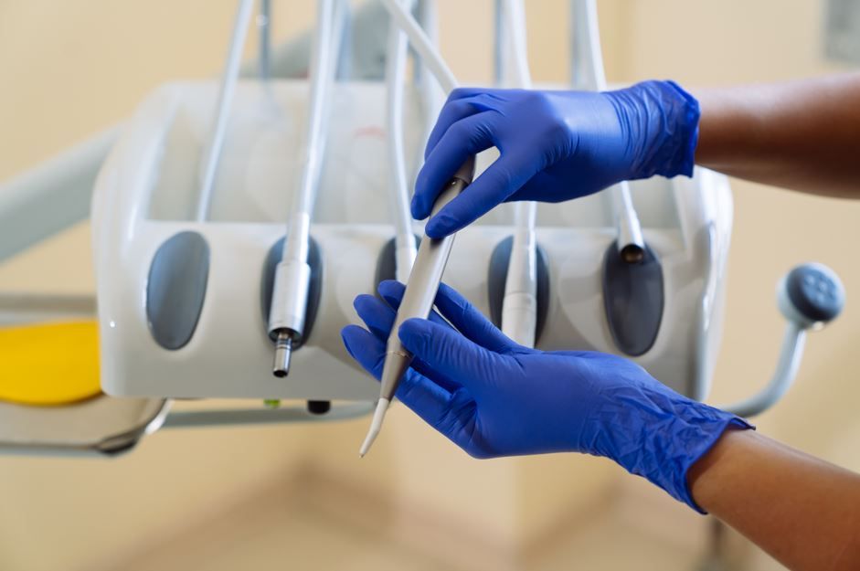 Manos con guantes azules sosteniendo herramienta dental, junto a equipo dental.