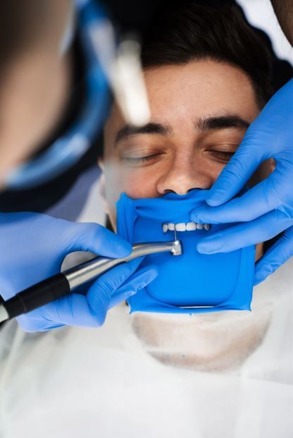 Persona sometida a trabajo dental; guantes azules y dique de goma; herramientas dentales en uso.