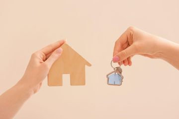 Manos sosteniendo una silueta de casa de madera y un pequeño llavero con forma de casa sobre un fondo beige.