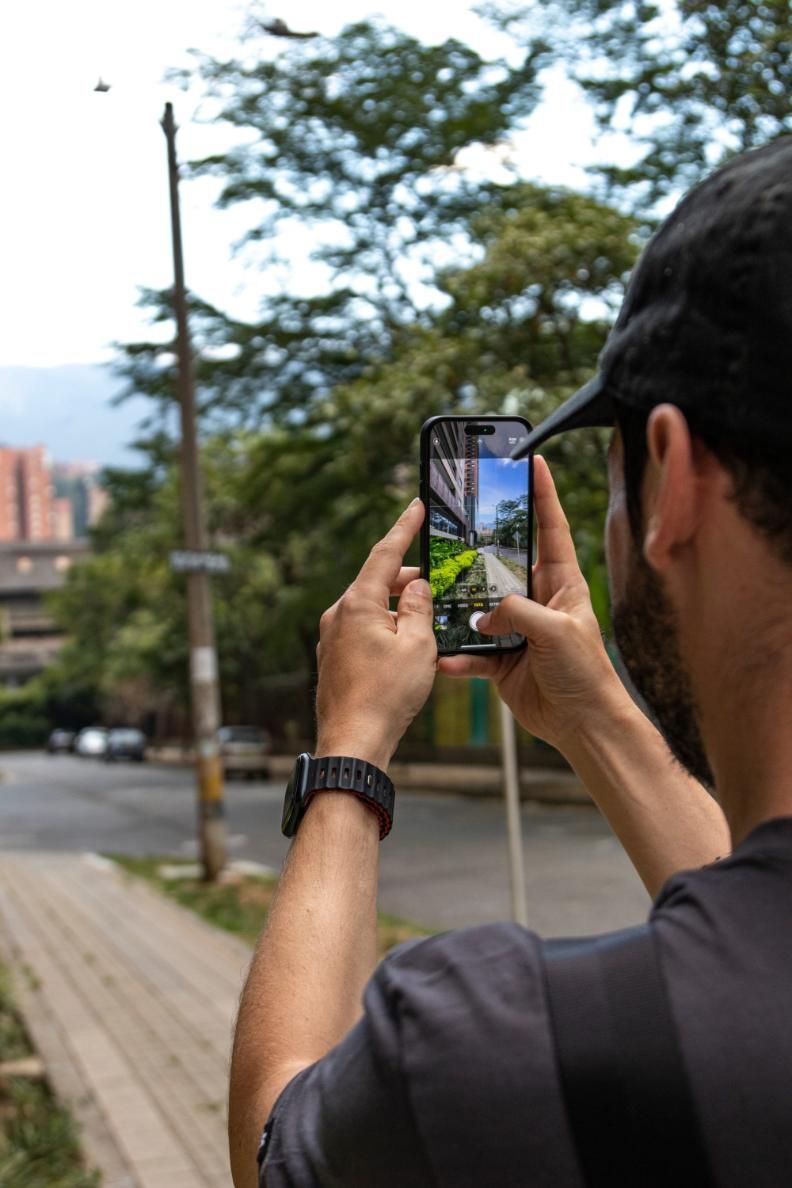 Una persona sostiene un teléfono para fotografiar una escena callejera con árboles y edificios.