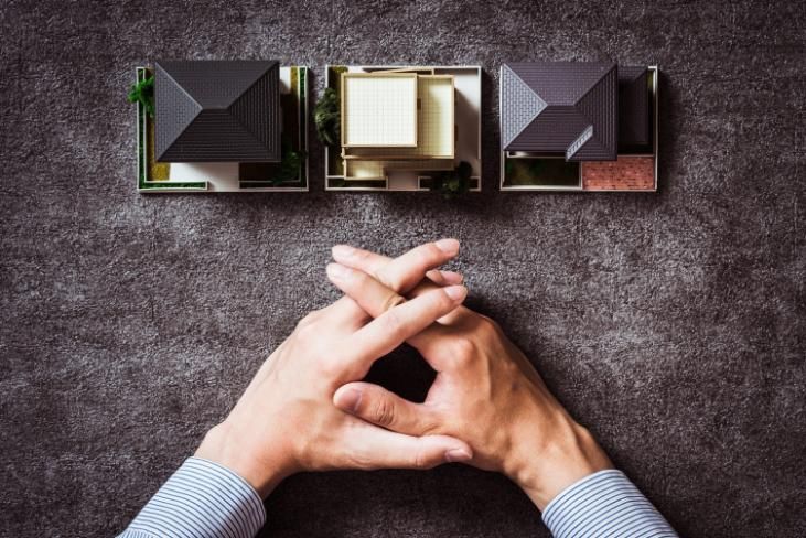Unas manos entrelazadas sobre un escritorio, debajo de tres maquetas de casas