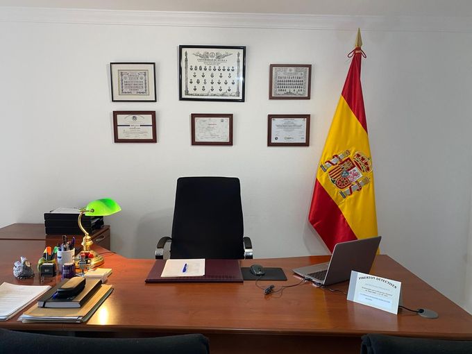 Escritorio de oficina con silla, certificados enmarcados en la pared y una bandera española en la esquina.