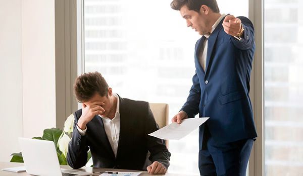 Dos oficinistas sentados en un escritorio; uno parece estresado mientras el otro está de pie sosteniendo papeles.