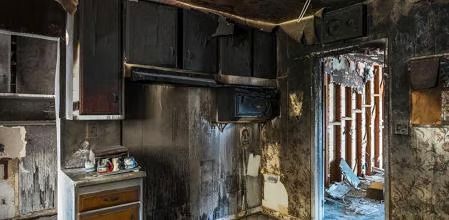 Cocina quemada y dañada por el humo, con armarios carbonizados y una puerta que da a la luz del día.