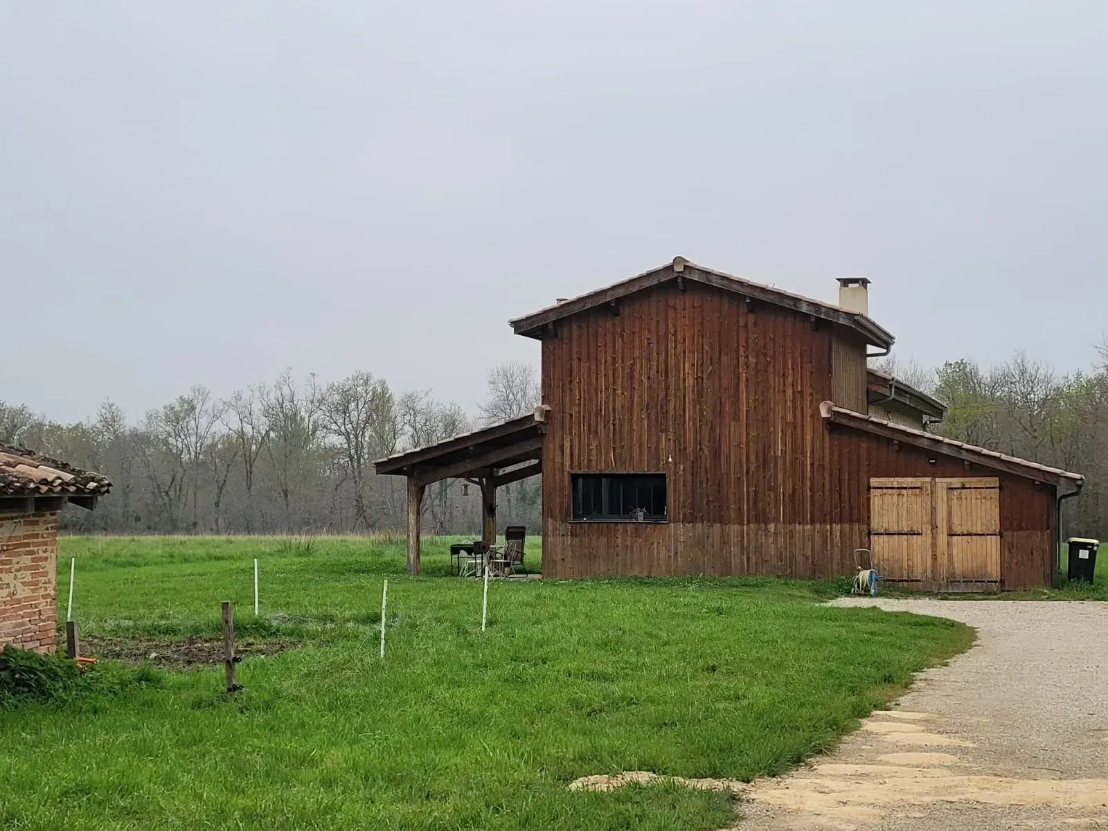 Bâtiment en bois de style grange avec porche couvert et petit jardin dans un champ herbeux. Ciel couvert.
