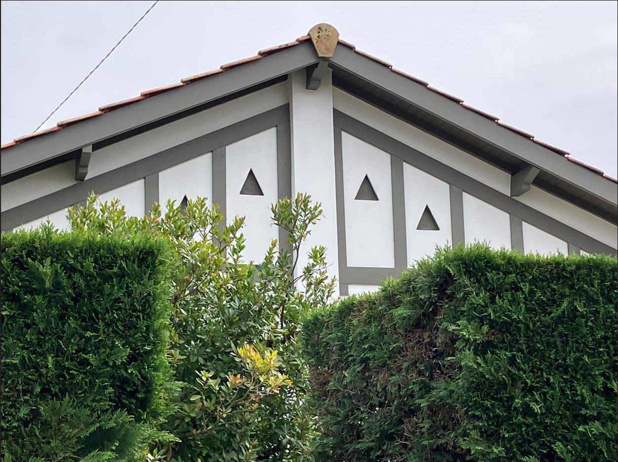Toit triangulaire d'un bâtiment blanc avec des finitions gris foncé, orné de petites découpes triangulaires et d'une haie.