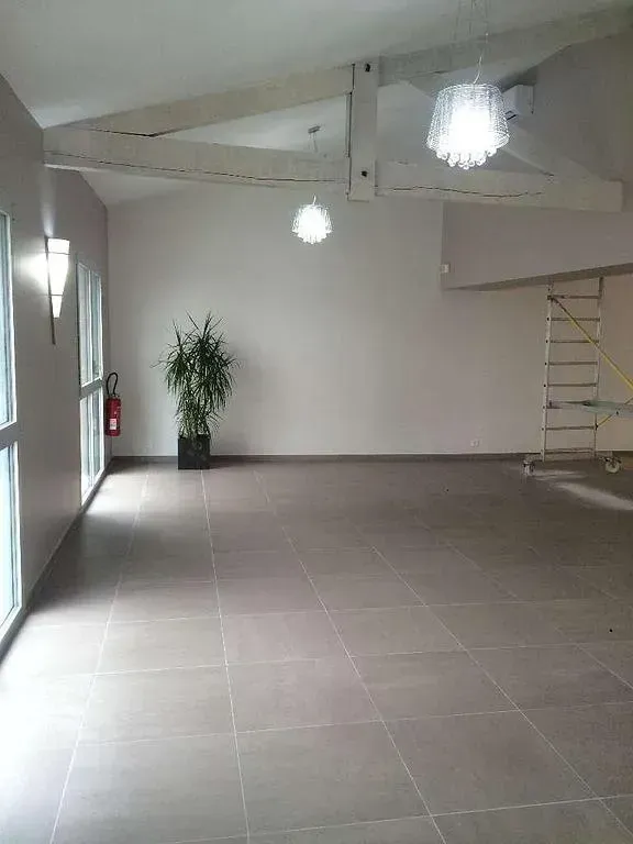 Pièce vide au sol carrelé clair, avec de hautes fenêtres et un éclairage décoratif. Une plante en pot se trouve au centre.