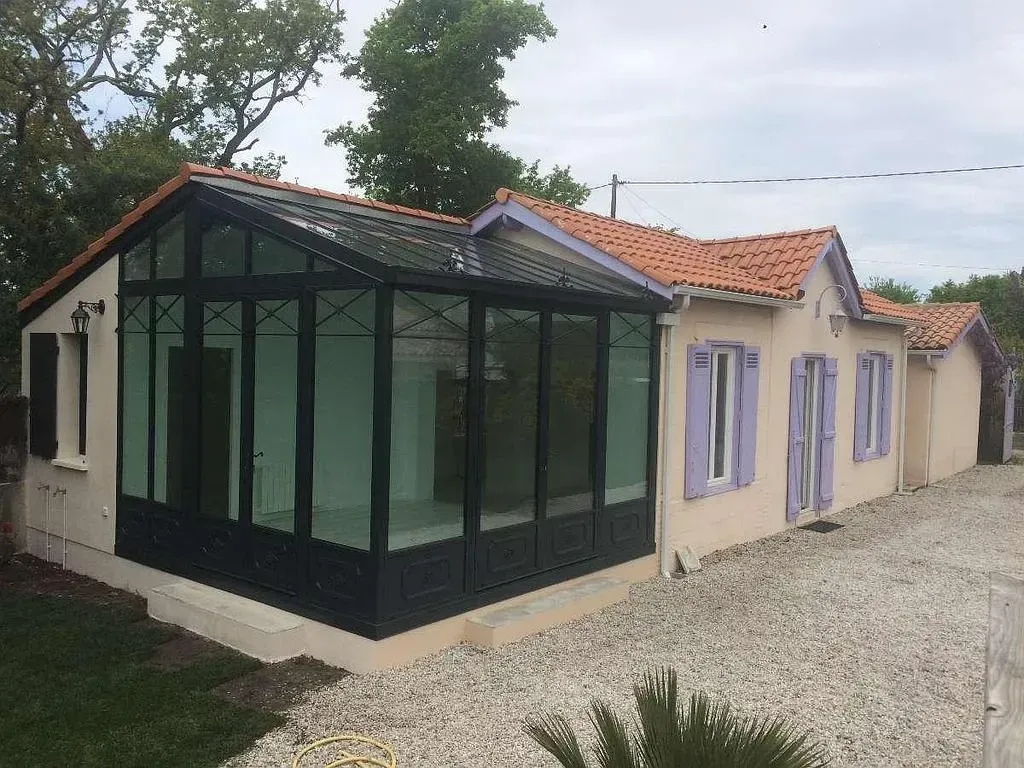 Maison beige avec une véranda en verre noir, des volets violets et des tuiles rouges.