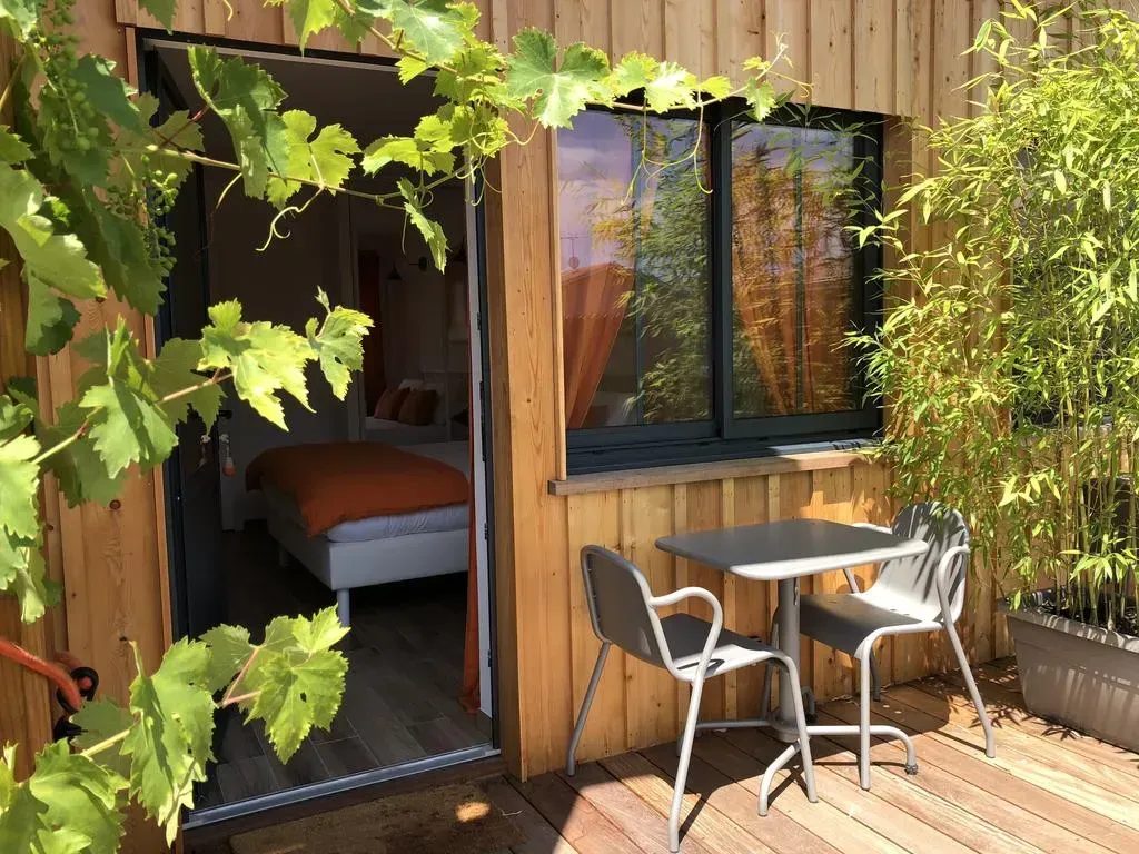 Extérieur d'une cabane en bois avec la porte ouverte d'une chambre. Une petite table et des chaises sont installées sur une terrasse. Des vignes encadrent l'ouverture.