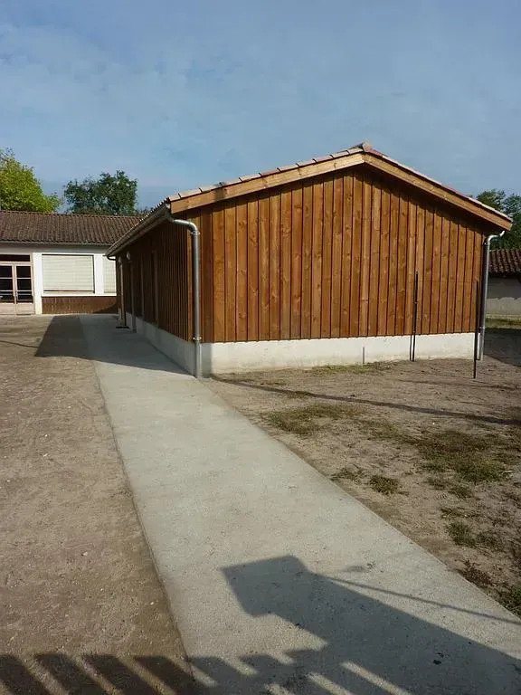 Bâtiment en bois avec chemin en béton, ciel gris et aperçu d'un deuxième bâtiment.