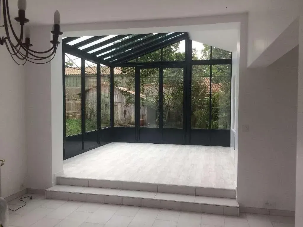 Vue intérieure d'une véranda avec parois vitrées, cadre noir et sol imitation bois. Donne sur un jardin.