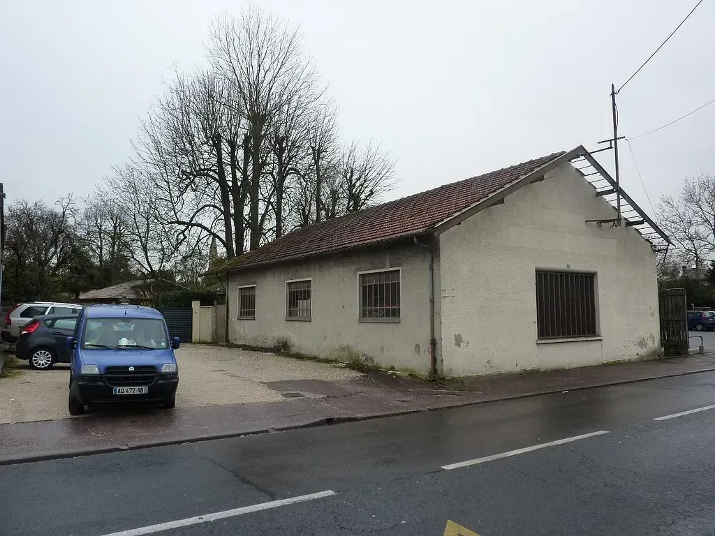 Petit bâtiment beige au toit de tuiles, véhicules garés devant, par une journée grise et nuageuse.