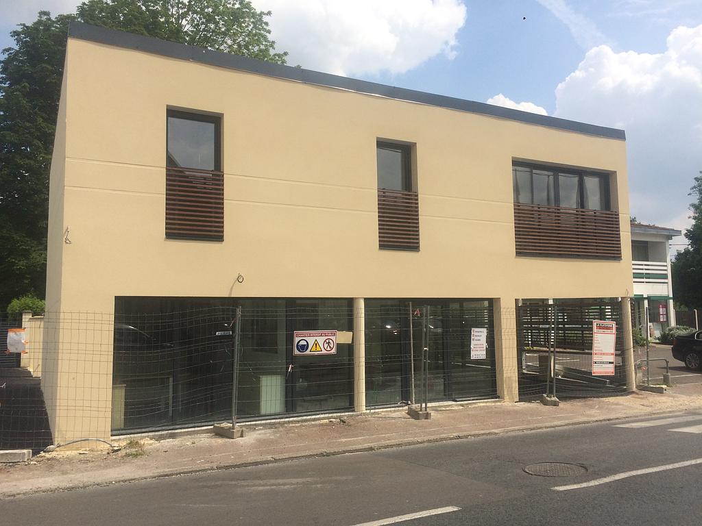 Bâtiment moderne de deux étages avec de grandes fenêtres au rez-de-chaussée, une façade beige et de petites fenêtres à l'étage avec des persiennes en bois.