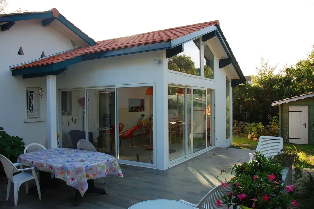 Maison blanche avec véranda vitrée et toit de tuiles rouges, coin repas extérieur avec jardin.
