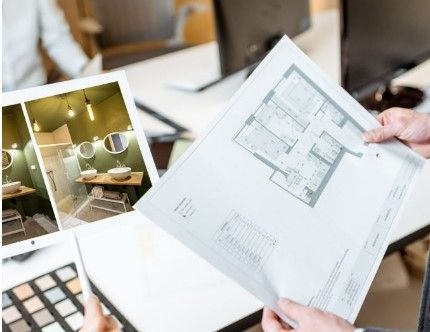 Des mains tenant un plan de bâtiment, avec un écran d'ordinateur affichant un plan de salle de bains et une palette de couleurs au premier plan.