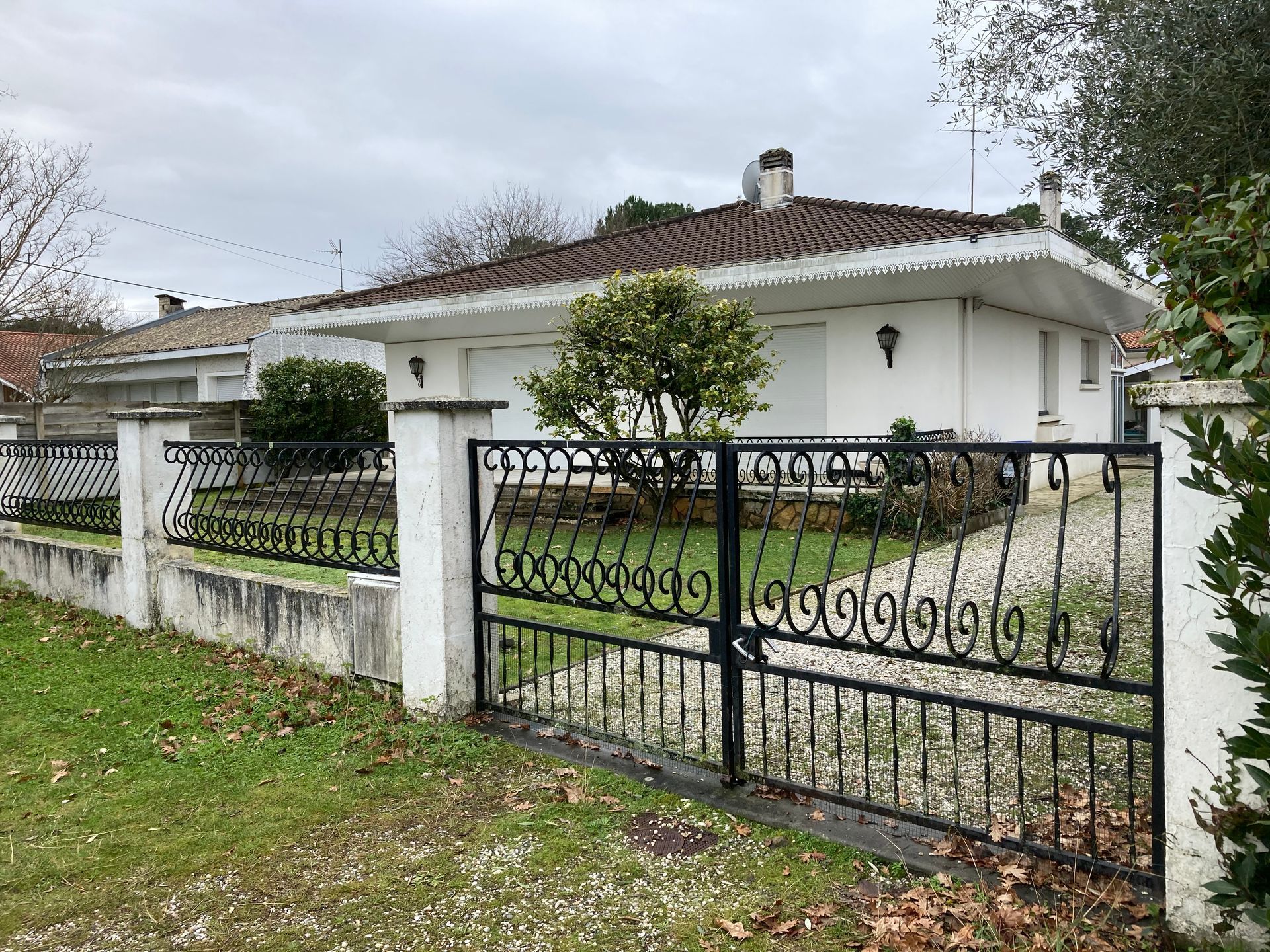 Maison blanche avec portail et clôture en fer forgé noir, entourée de pelouse et d'arbres.